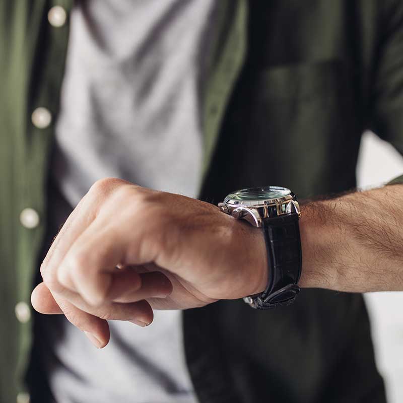 Mand med grøn skjorte udover hvis t-shirt kigger på sit armbåndsur for at se, hvad klokken er.